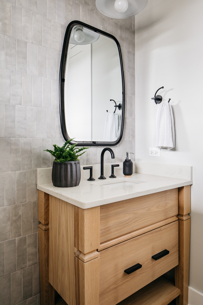 bathroom with light gray tiled walls and a brown wood vanity. White countertop and dark metallic decor.