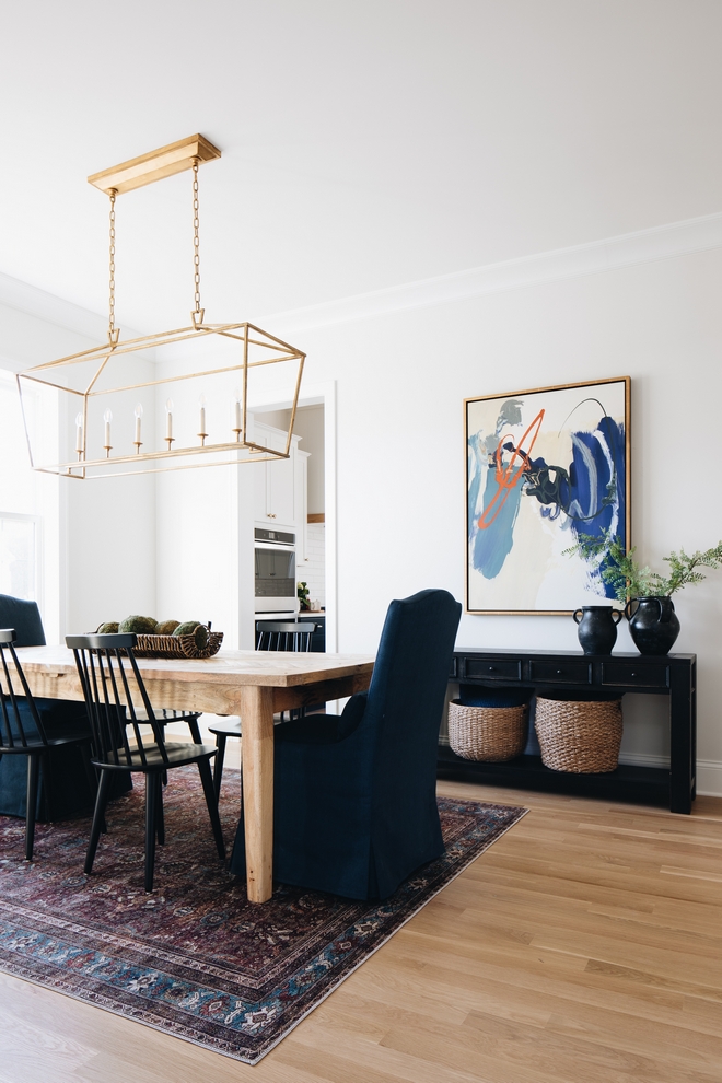 Dining room with dark rug, light wood table, dark chairs and a rectangular lantern chandelier. navy accents are found in the art, vases, chair, and rug.