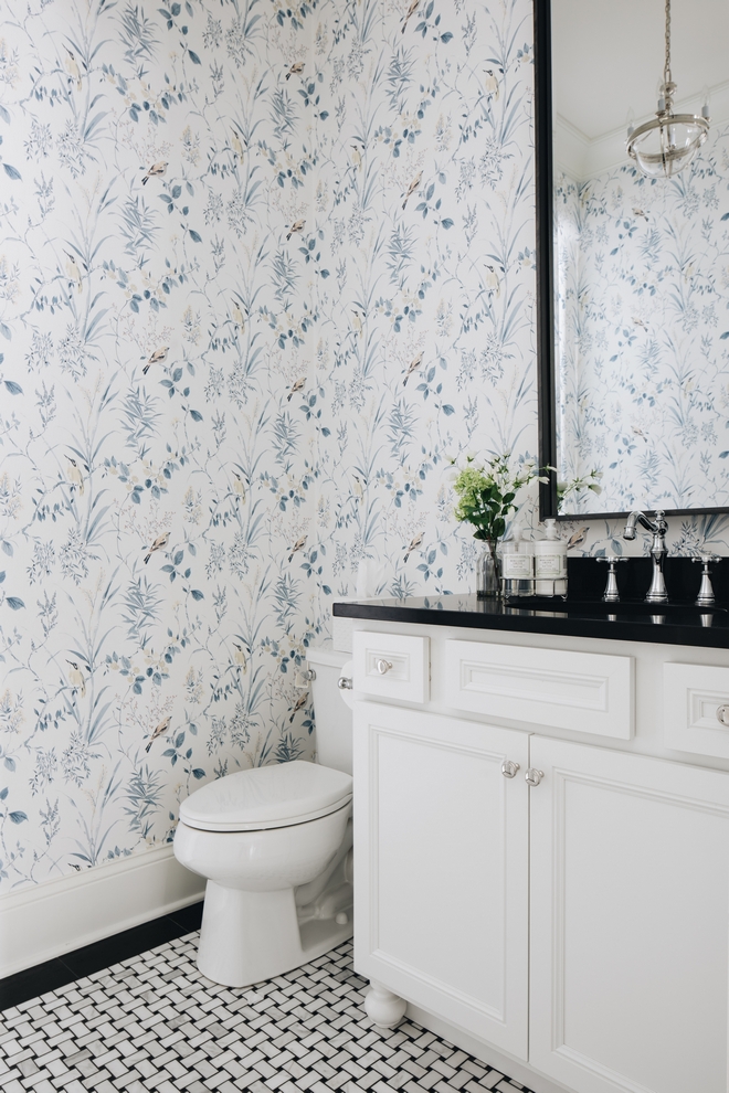 Powder bathroom with white and blue flowered southern wallpaper and black and white woven looking tile flooring. White vanity with black counter.