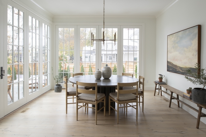 European style dining room with dark refurbished wood round table, french doors on one side and wall to wall to ceiling windows on another. Last wall has a large landscape picture with a long rustic decorative bench underneath