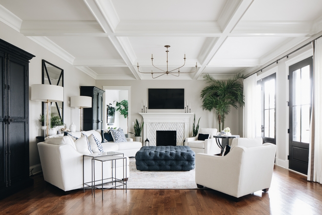 Great room with a light rug over wooden flooring. Black window doors on one wall with a blue ottoman in the center of the room surrounded by white sofa and white armchairs. 