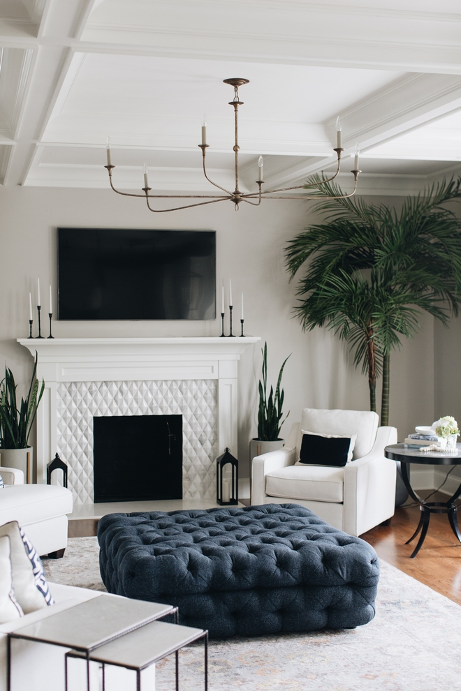 Closer view of great room. Large blue tufted ottoman is shown in front of a white armchair and white tiled fireplace. Decorative southern style plants are in the corner and on either side of the fireplace