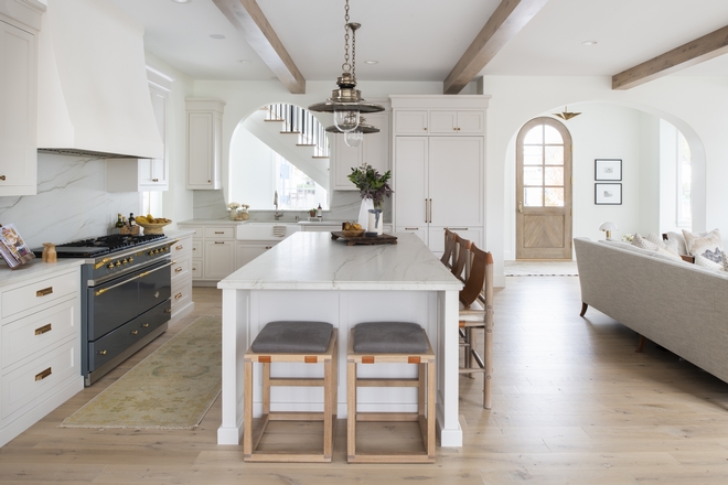 Side view of kitchen with backless barstools with leather accents, exposed bulb pendant lights, white cabinets and counters, brass accents and brass pulls, exposed beam ceiling, disguised fridge, and arch in front of kitchen sink.