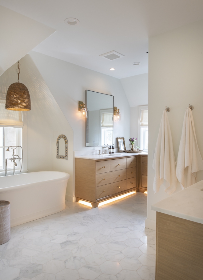 Master bathroom with a free standing bathtub in a nook with a window and overhanging woven bell shaped pendant. Pale wood vanity with recessed lighting under - single mirror with simple glass sconces and a white and gray marble hexagonal tile floor