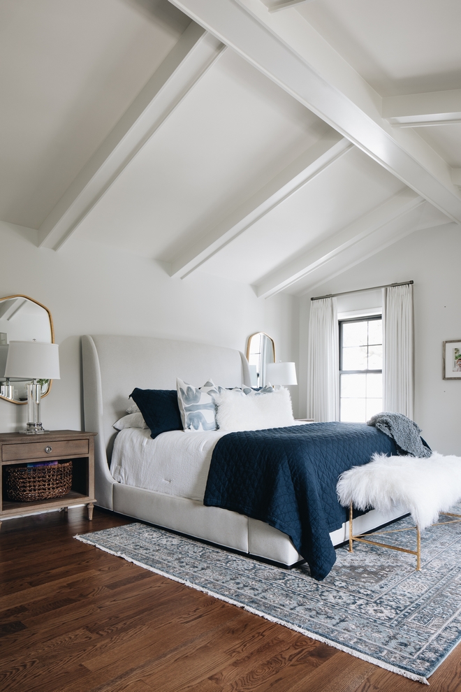 Master bedroom with large white bed frame, faux fur bench, dark blue accent pillow and blanket, side tables, vaulted ceiling, and accent area rug.