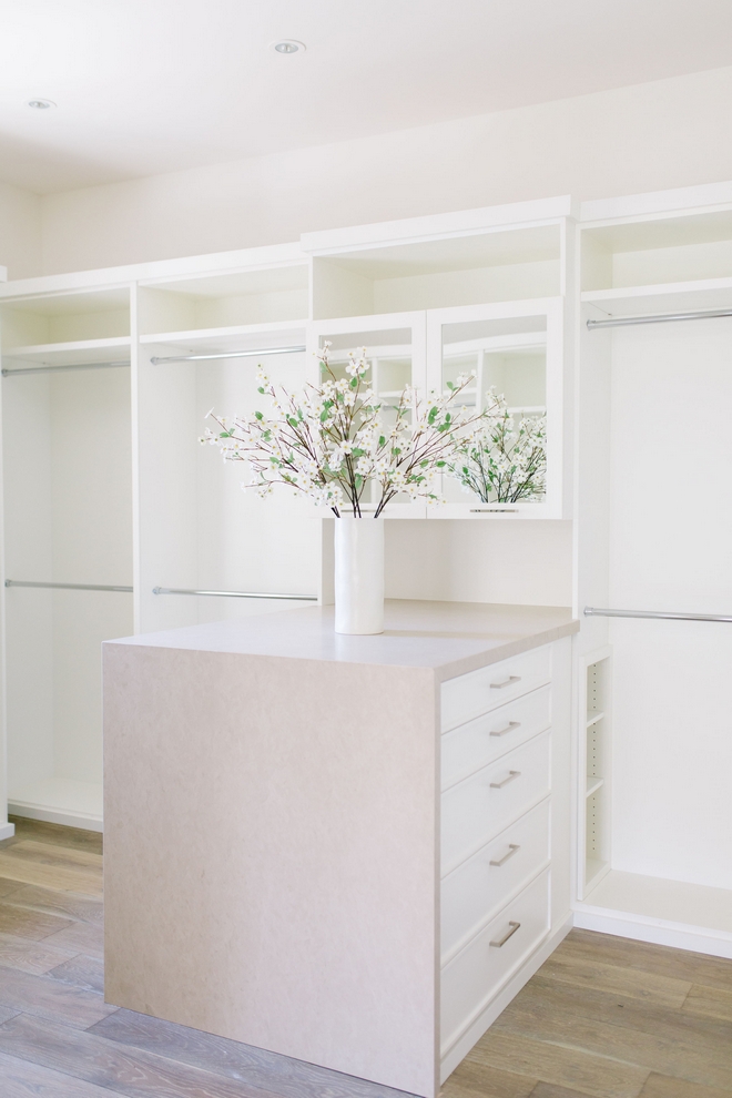 closet with white two-level shelves and rods and natural wood flooring. A square peninsula with drawers is the center of the room.