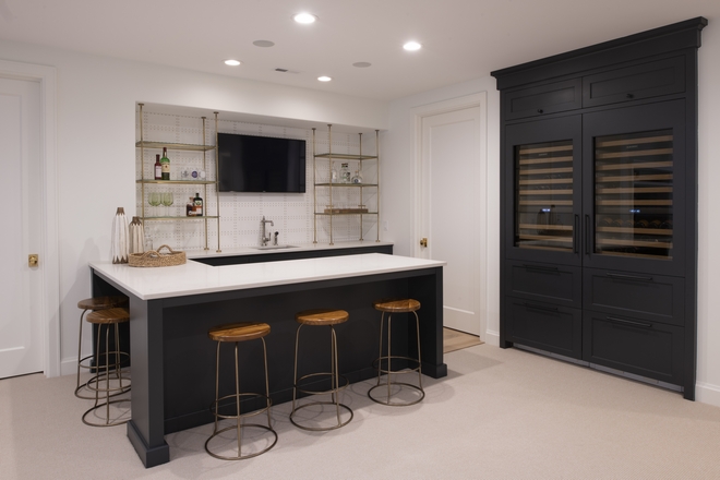 European style basement bar area with dark gray island and wine cabinet, pale short carpet, white wall, backsplash, and counter with brass accents and backless round counter stools on two sides